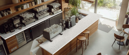 Modern coffee shop with MSI Iced White Polished Quartz countertop, espresso machines, wood shelves, large windows, and bright decor.